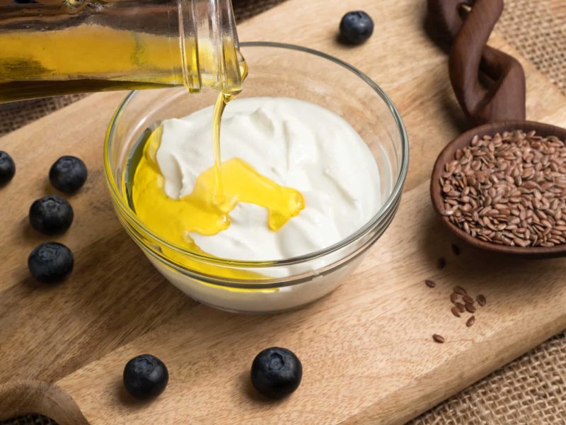 Flax seed oil is being poured over cottage cheese in a bowl, with flax seeds and blueberries in the background